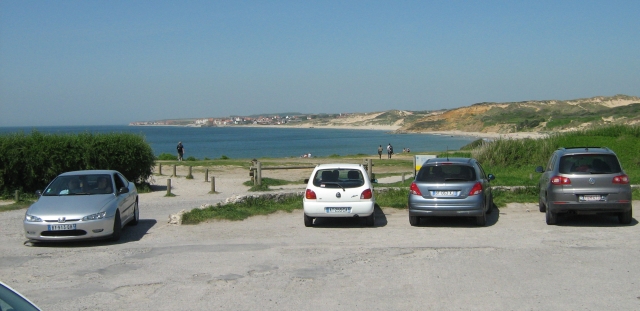 inf the foreground cars on a gravel carpark overlooking a long curving sandy shoreline with dunes